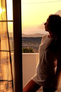 Side view of woman looking through window at sunset