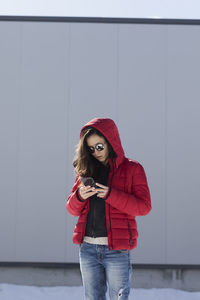 Young woman using phone while standing against wall