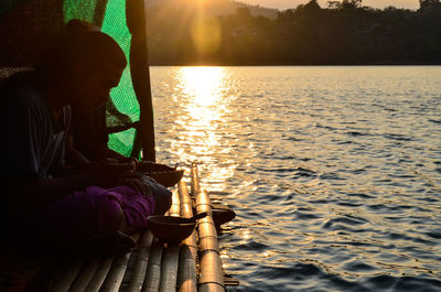 Woman sitting by lake against sky during sunset