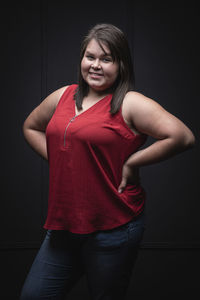 Charming self assured young overweight female wearing casual red blouse and pants keeping hands on waist and looking at camera with smile on black background