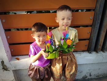 Cute brothers wearing traditional clothing holding flowers against wall