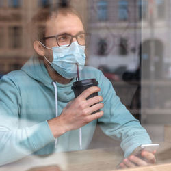 Man wearing mask drinking coffee in cafe seen through window