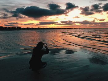 Silhouette woman photographing on beach against sky during sunset