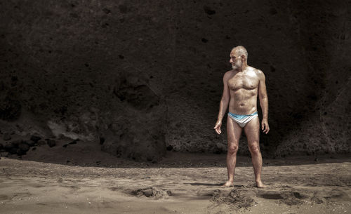 Portrait of shirtless man in swimwear standing on beach against rock 