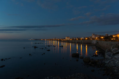 Illuminated city by sea against sky at sunset