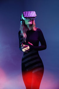 Young unrecognizable blonde woman standing wearing virtual reality headset on colorful blue neon illumination studio background