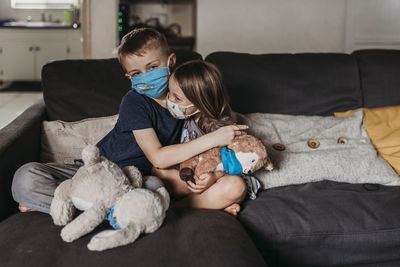 Young girl and school-age boy with masks hugging and smiling on couch