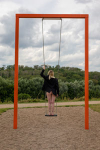 Rear view of woman standing on swing against sky