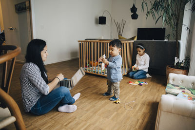 Mother using laptop while working from home sitting with children in living room