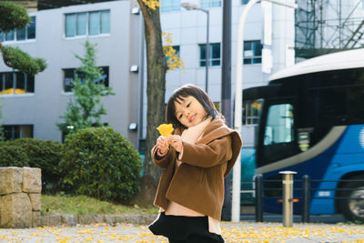 Girl holding yellow leaves while standing against buildings