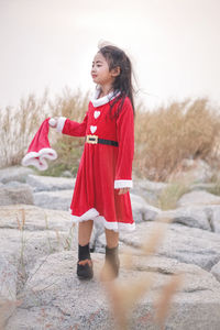 Girl wearing red dress holding santa hat while standing on rocks against sky