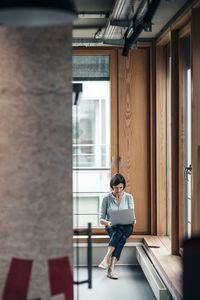 Female entrepreneur working on laptop while sitting over alcove window seat at office