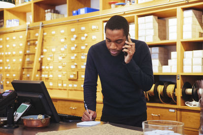 Young male owner talking on smart phone while writing at checkout counter in furniture store