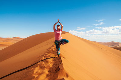 Woman practicing yoga on sand dune in desert against sky