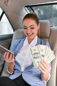 Smiling businesswoman with paper currency talking on phone while sitting in car