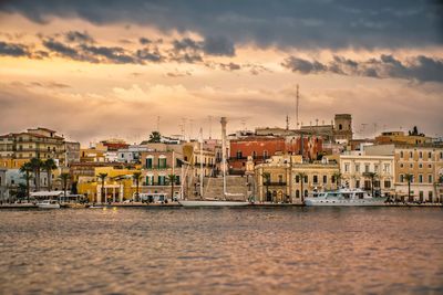 Sailboats in sea by buildings against sky during sunset