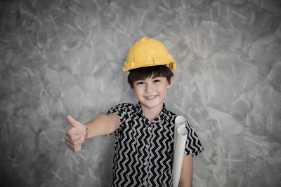 Portrait of smiling boy wearing hardhat gesturing thumbs up against wall