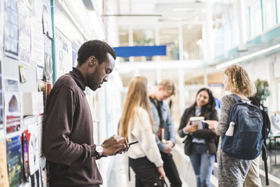 Side view of young male student using mobile phone while leaning on wall at university