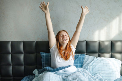 Full length of woman with arms raised sitting on bed at home