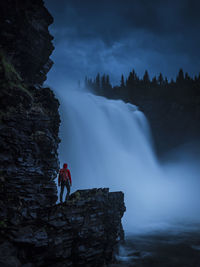 Hiker looking at tannforsen waterfall while standing on rocks against sky