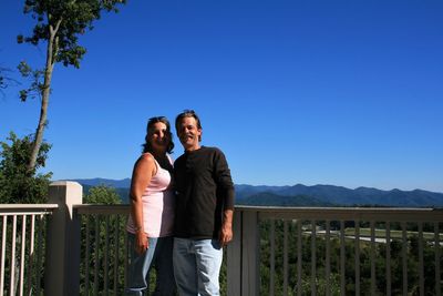 Portrait of smiling couple standing at balcony against clear blue sky