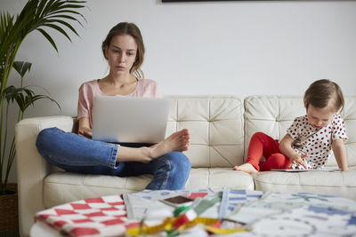 Fashion designer looking at laptop while daughter using digital tablet on sofa at home