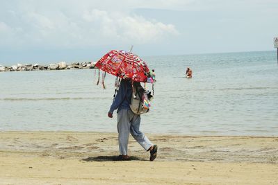 Side view of man with colorful umbrella walking at beach against sky on sunny day