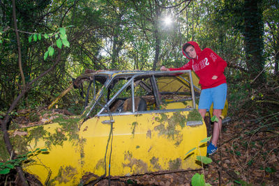 Boy standing on old yellow abandoned car in forest