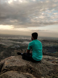 Rear view of man sitting on rock by sea against sky