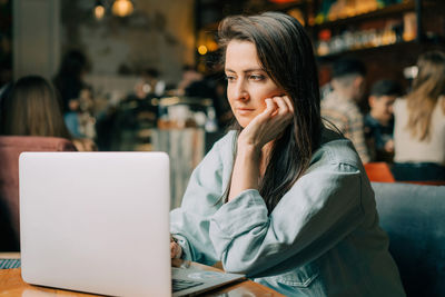 A young brunette woman sits in a cafe and works on a laptop.