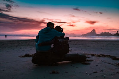 Man sitting on shore at beach against sky during sunset