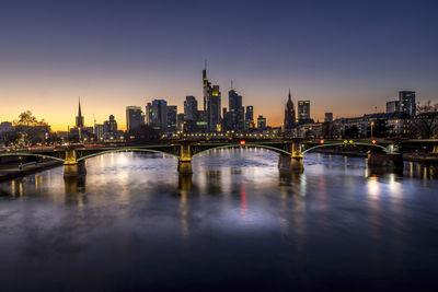 Illuminated bridge over river by buildings against sky in city