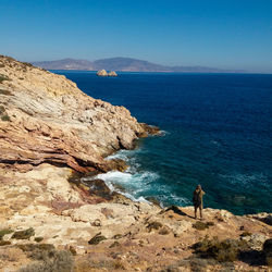 Rear view of man standing on rock formation by sea against blue sky