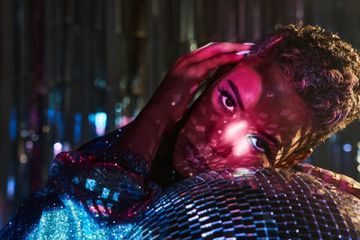 Trendy young ethnic female touching face while looking at camera and leaning on mirror ball in nightclub