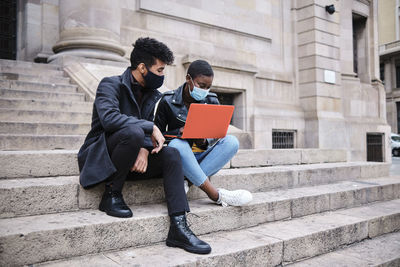 Friends wearing protective face mask using laptop while sitting on steps in city