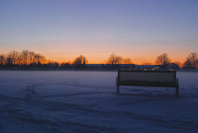 Empty bench on snow field against sky during sunset