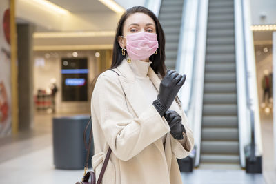 Beautiful woman in pink medical mask putting on leather gloves in a public place