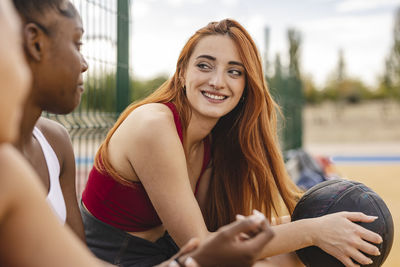 Smiling young woman holding basketball sitting with friends in basketball court