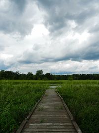 Empty walkway leading towards grassy field against cloudy sky