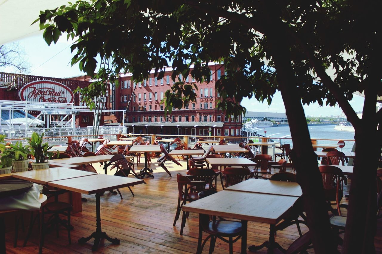 table, chair, restaurant, tree, built structure, architecture, water, absence, building exterior, sidewalk cafe, sky, tourist resort, potted plant, day, empty, sunlight, sea, wood - material, palm tree, place setting