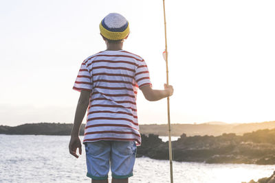 Rear view of boy holding fishing rod while standing against sky