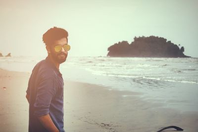 Portrait of young woman wearing sunglasses standing on beach against clear sky