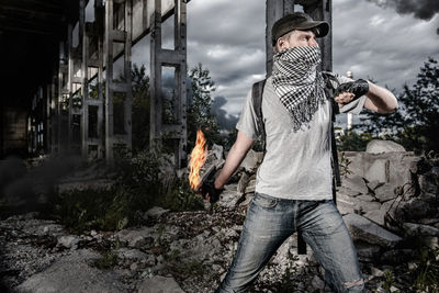 Young man wearig mask throwing bottle while fire under bridge