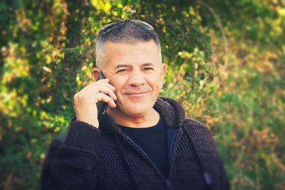 Portrait of smiling mature man talking on mobile phone while standing against plants