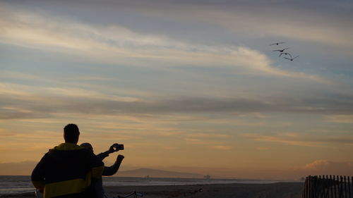 Rear view of man photographing sea against sky during sunset