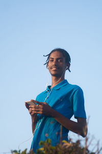 Low angle view of young adult standing against clear sky holding a mobile phone