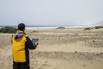 Rear view of man using digital tablet while standing at beach against clear sky