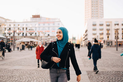 Smiling young muslim woman wearing hijab walking on footpath in city