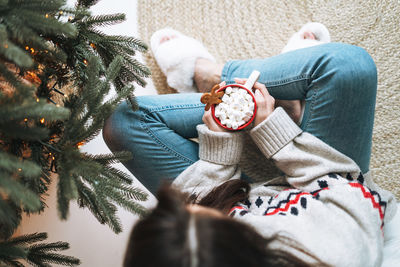 Young woman in sweater with cup of cocoa with marshmallow in room with christmas tree at home