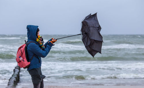 Woman holding damaged umbrella while standing at shore of beach against sky